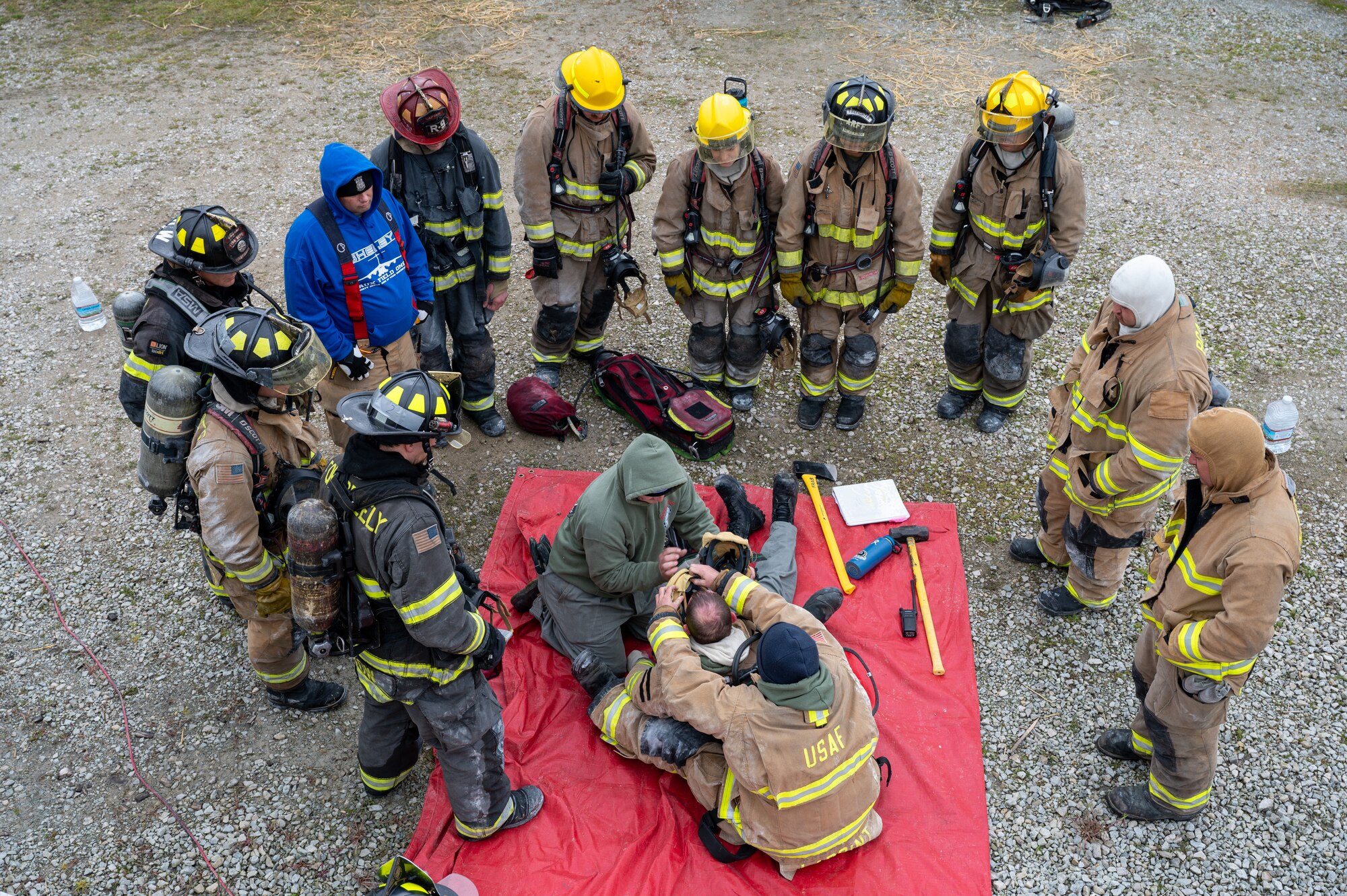 An aerial shot of a group of firefighters standing in a circle, looking down at a gear demonstration by other firefighters on the ground.