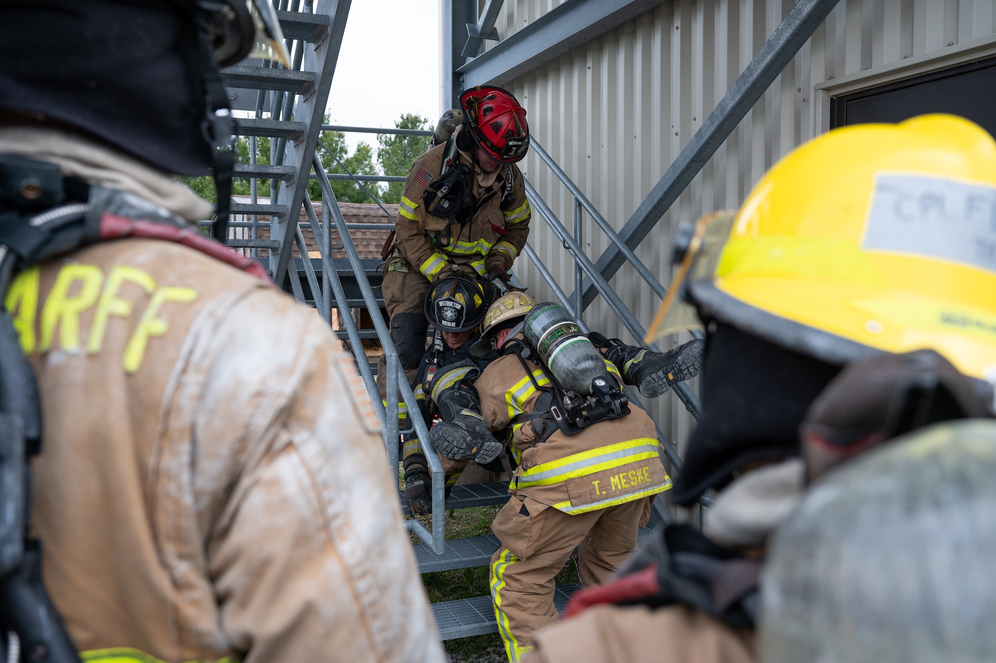 Firefighters in the foreground watch as firefighters in the background carry another firefighter, wearing a hat that says "Instructor", up a flight of stairs. The front three firefighters are instructors and the back two are students.