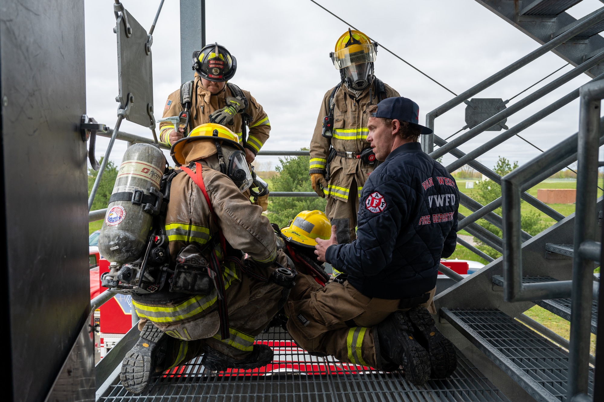 A group of firefighters in a circle. All in uniform except one at front right who is in a bomber-style jacket and baseball cap.