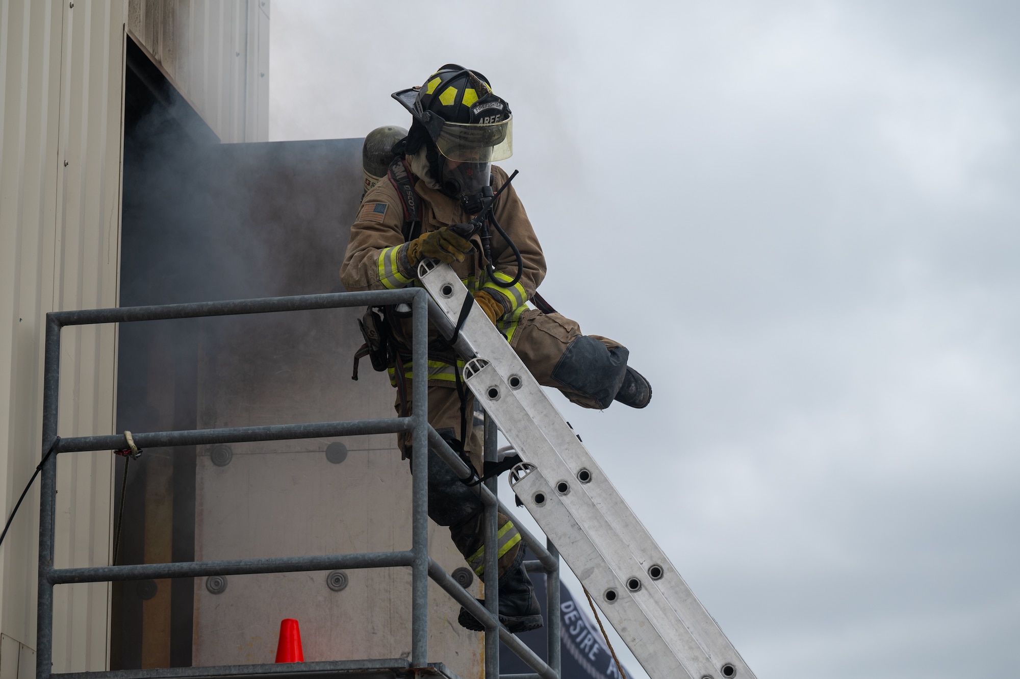 A firefighter in full protective gear swings his leg over a bar. On the other side of the bar, leaned against the platform he was once standing on, a ladder.