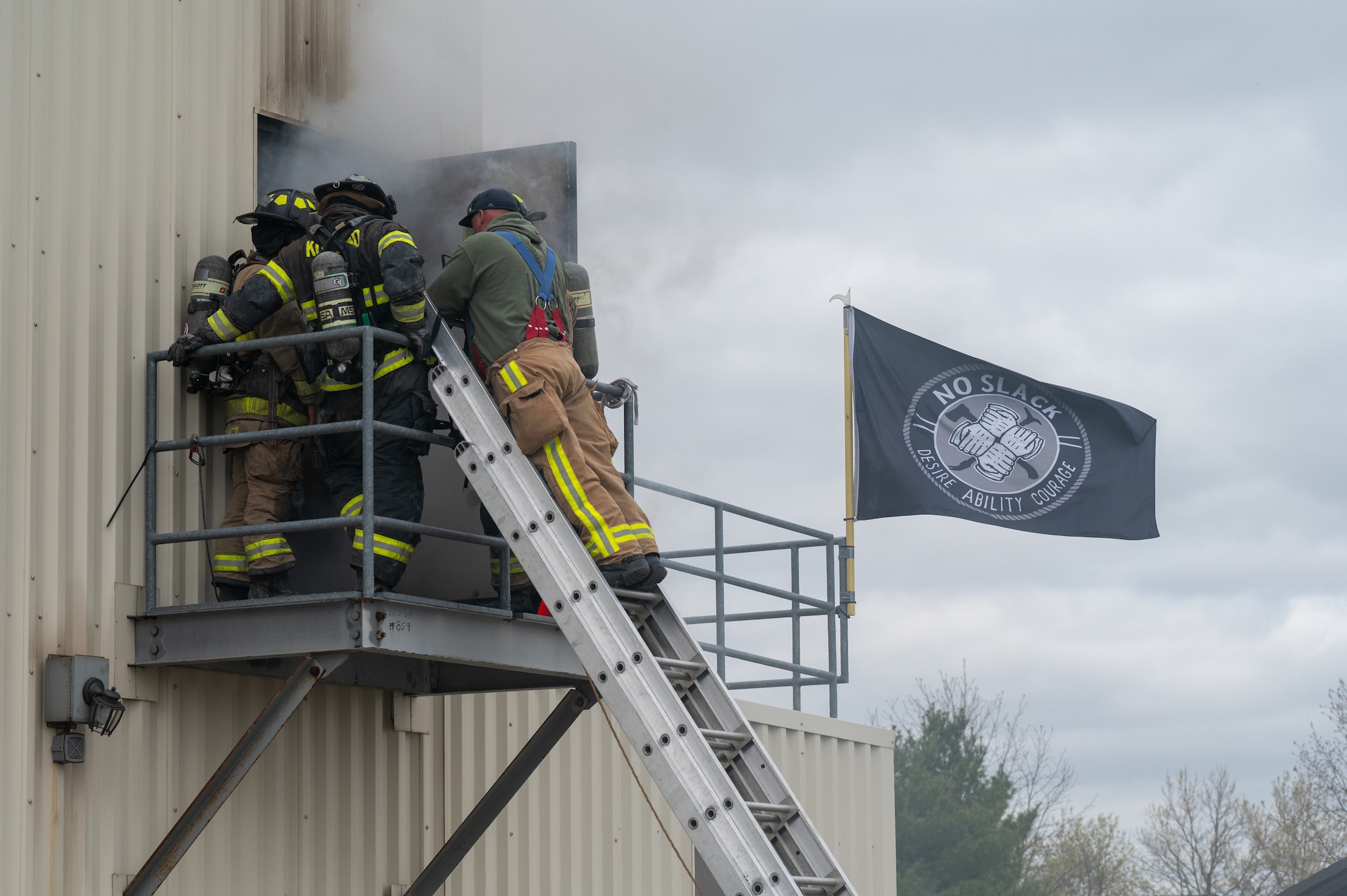 A man atop a leaned ladder talks to a group of firefighters on the platform that he is now level with. In the background, a black flag that reads "NO SLACK".