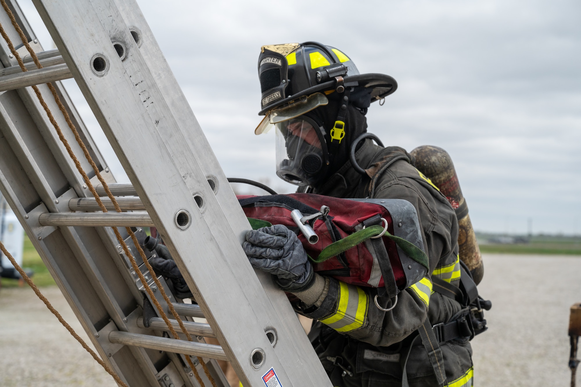A firefighter ascends a metal ladder holding a red bag.