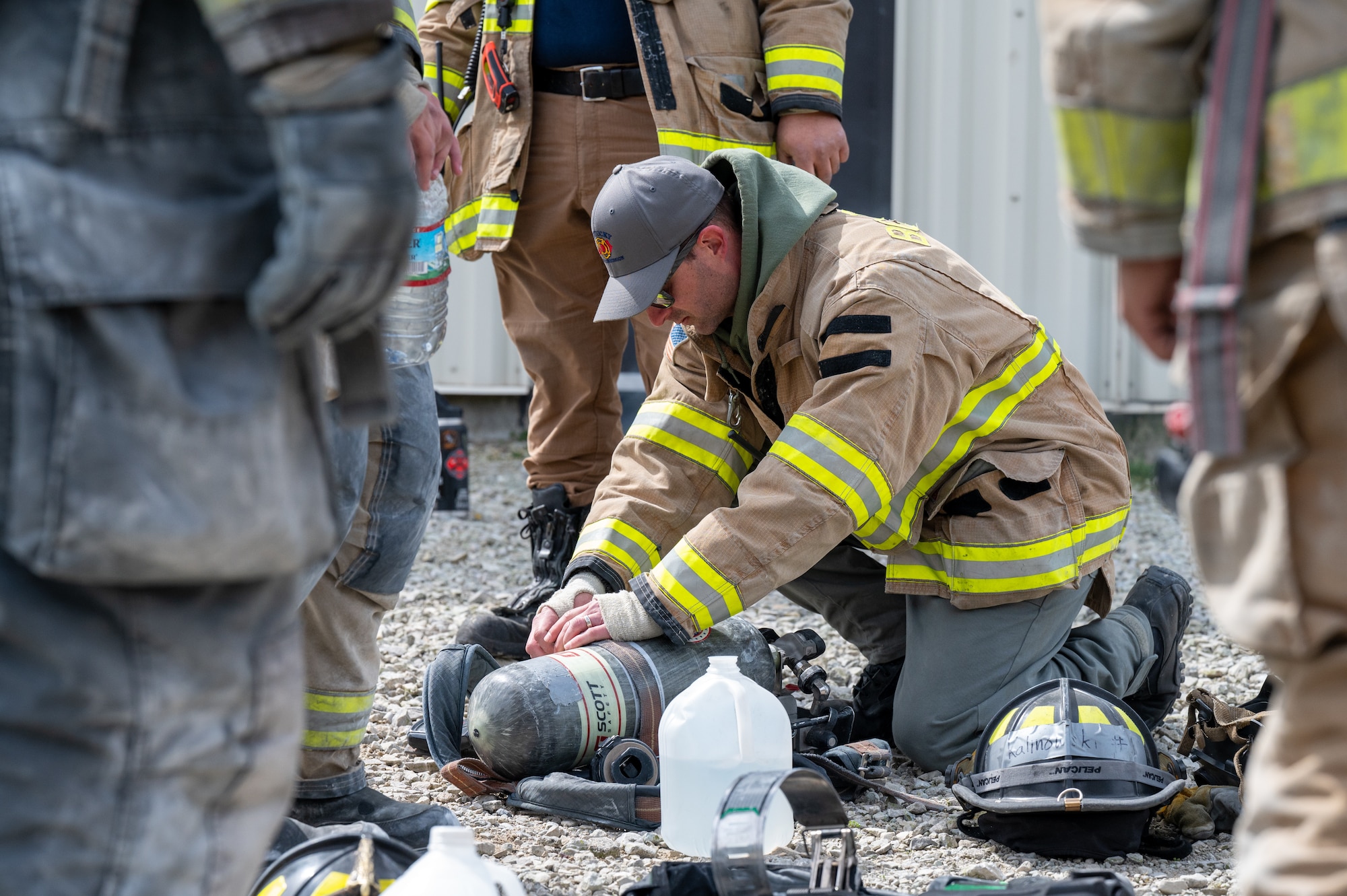 A man with both knees on the ground, leaning over a firefighter's oxygen tank and making adjustments to it with his hands.