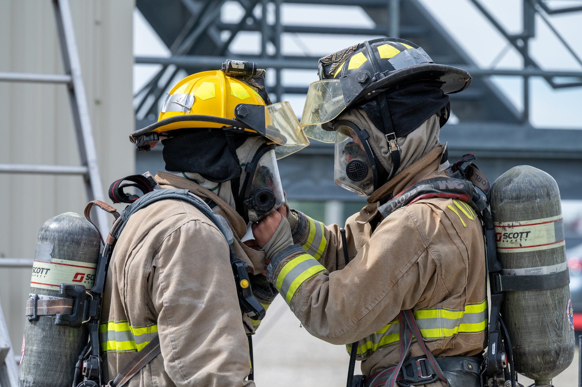 Two firefighters, facing face to face. One adjusts the other's mask. Both are wearing full protective gear.