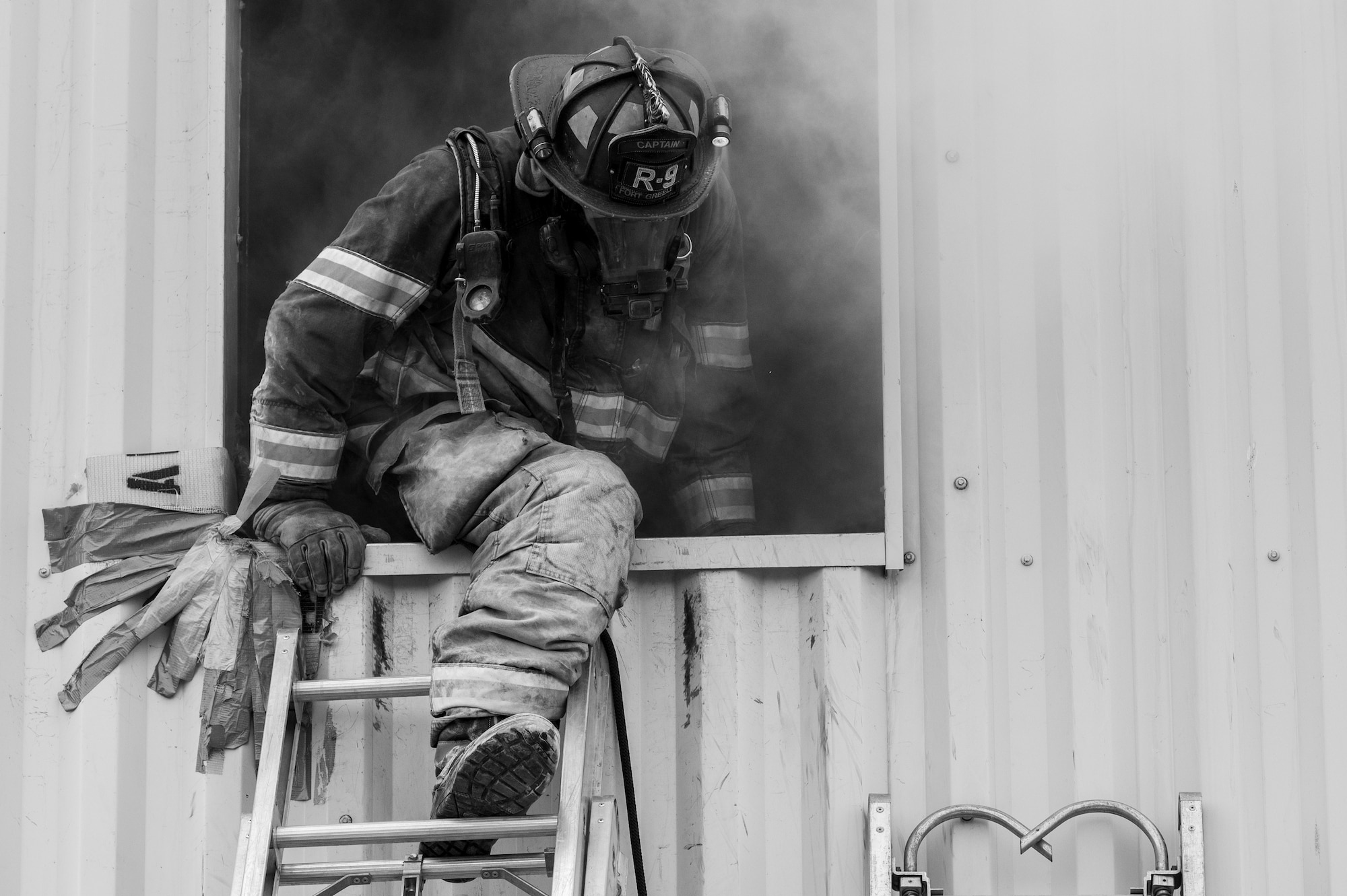 A masked firefighter exits a smoking building via window. Leaned up against the window, a metal ladder is visible.