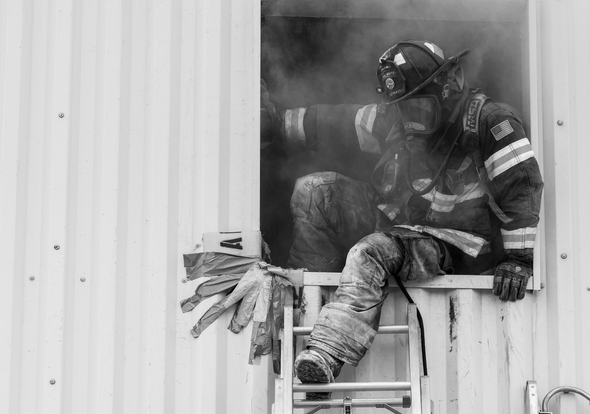 A masked firefighter exits a window via ladder. He is sitting in the frame of the window, with his foot on the second top most rung of the ladder.