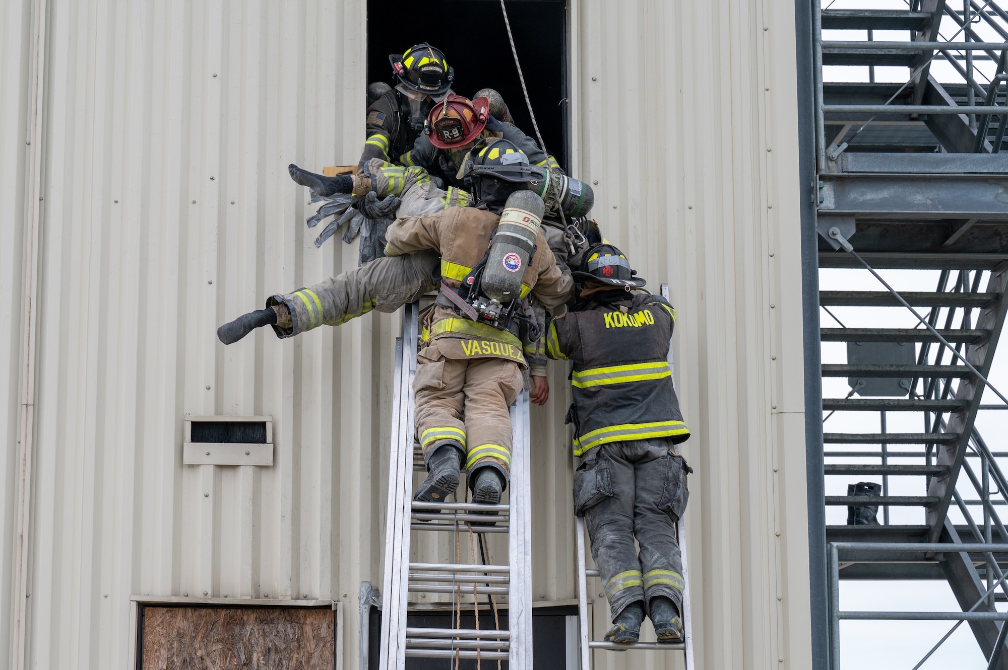 Two firefighters stand on two ladders, side by side. At the top of the ladder, two firefighters lifting another firefighter out of the window and down to the laddered firefighters.