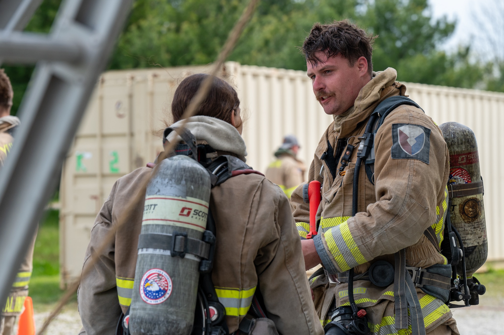 Two firefighters, in uniform, having a conversation. In the background, a shipping container. Framing the photo at left, a glimpse of a ladder and rope.