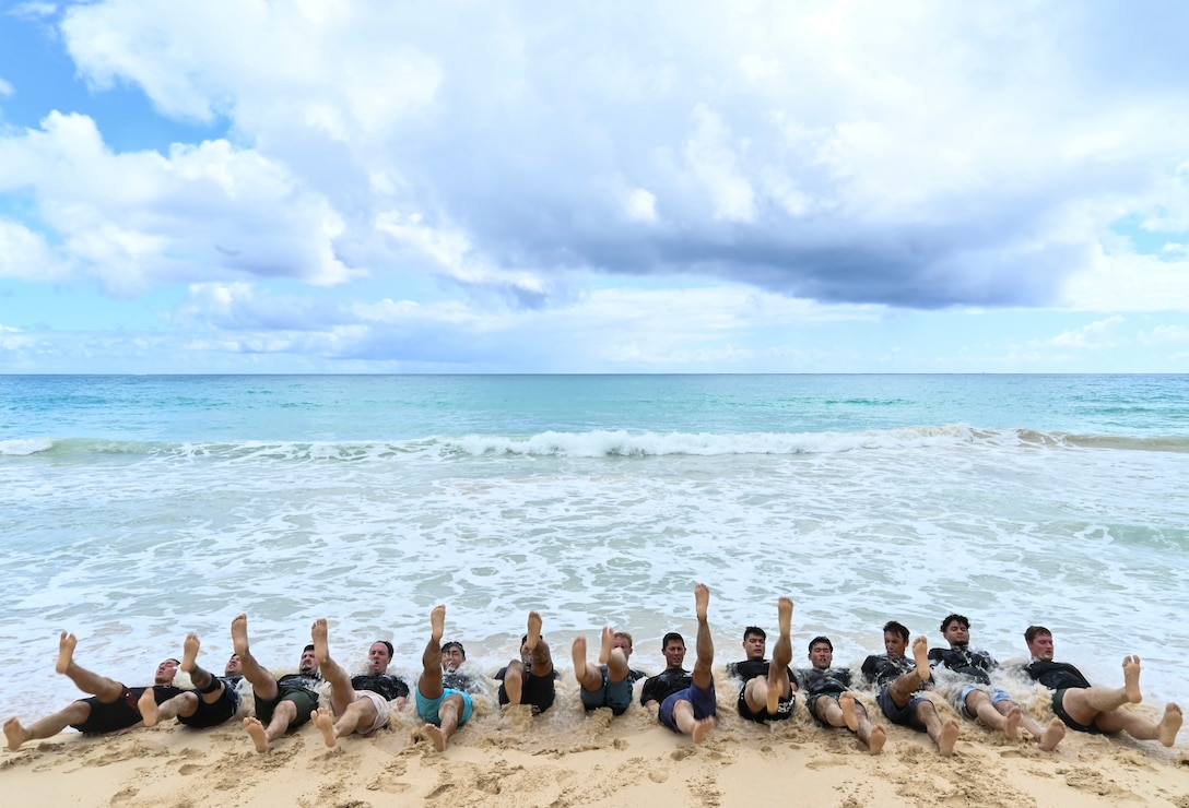 Recruits lie in a row doing exercises on their backs on a sandy beach.