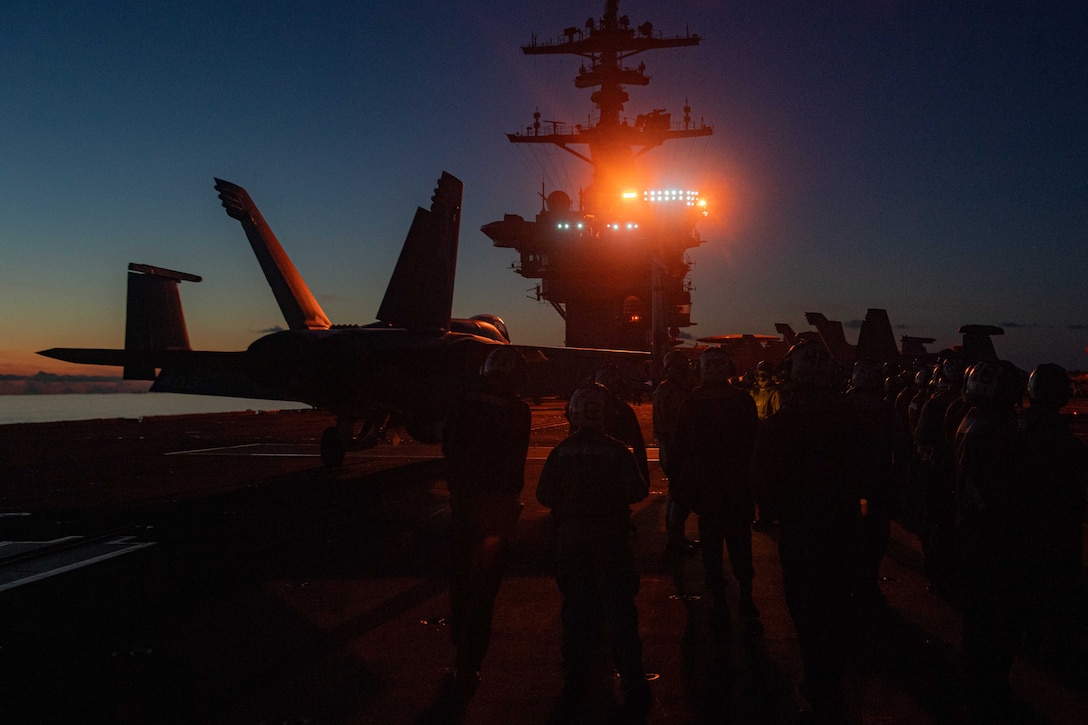 Sailors stand around a military aircraft on a ship lit by a small array of bright lights.