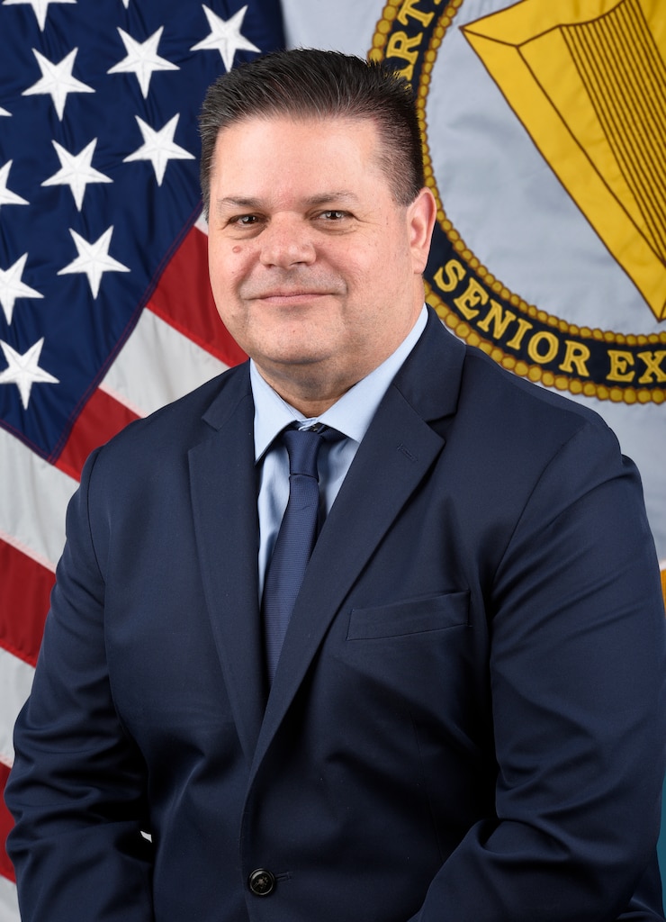 A man in a suit poses for a photo in front of the American and Senior Executive Service flags