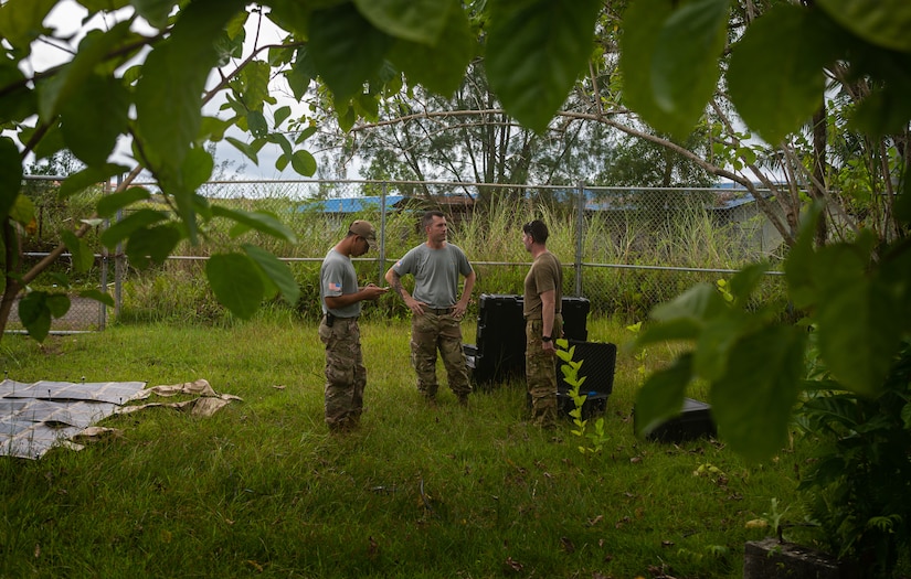 Three men in military uniform talk while at an excavation site.