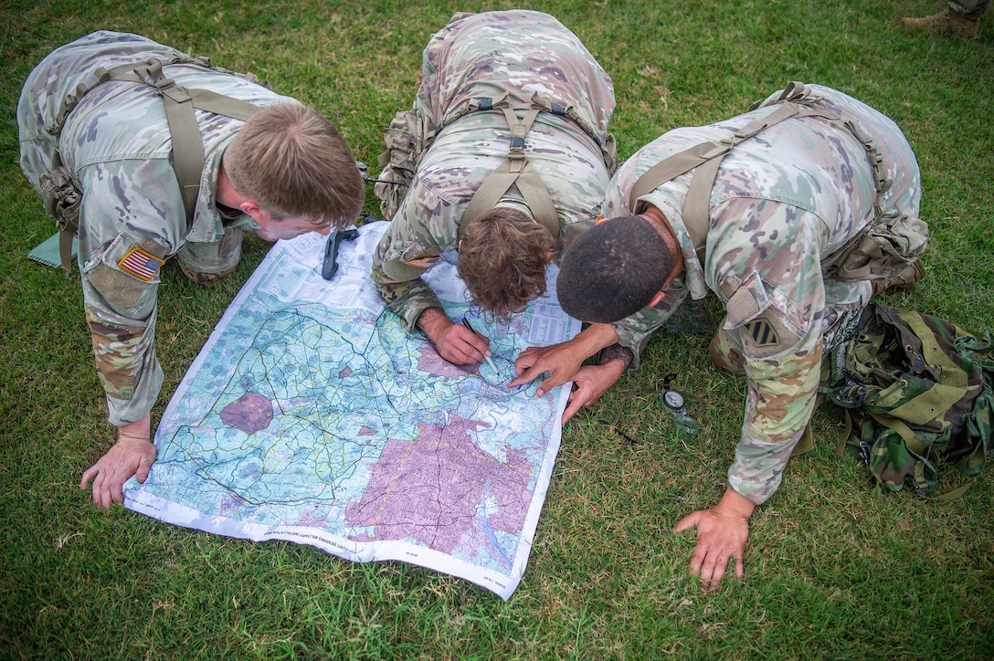 Three soldiers seen from above kneel over a map spread out in a grassy field.
