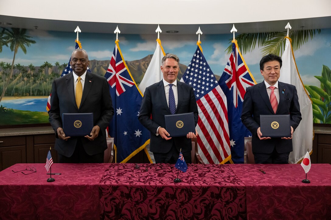 Secretary of Defense Lloyd J. Austin III stands at a table with his Australian and Japanese counterparts in front of the three nations' flags.