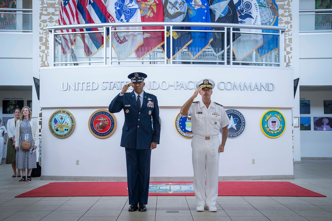 Air Force Gen. CQ Brown, Jr., salutes alongside a Navy admiral in a lobby-type space.
