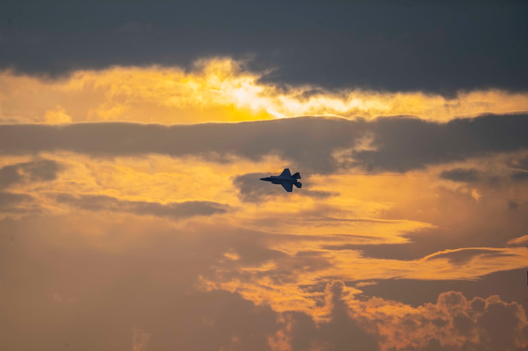 A military jet flies across an orange partially cloudy sky.