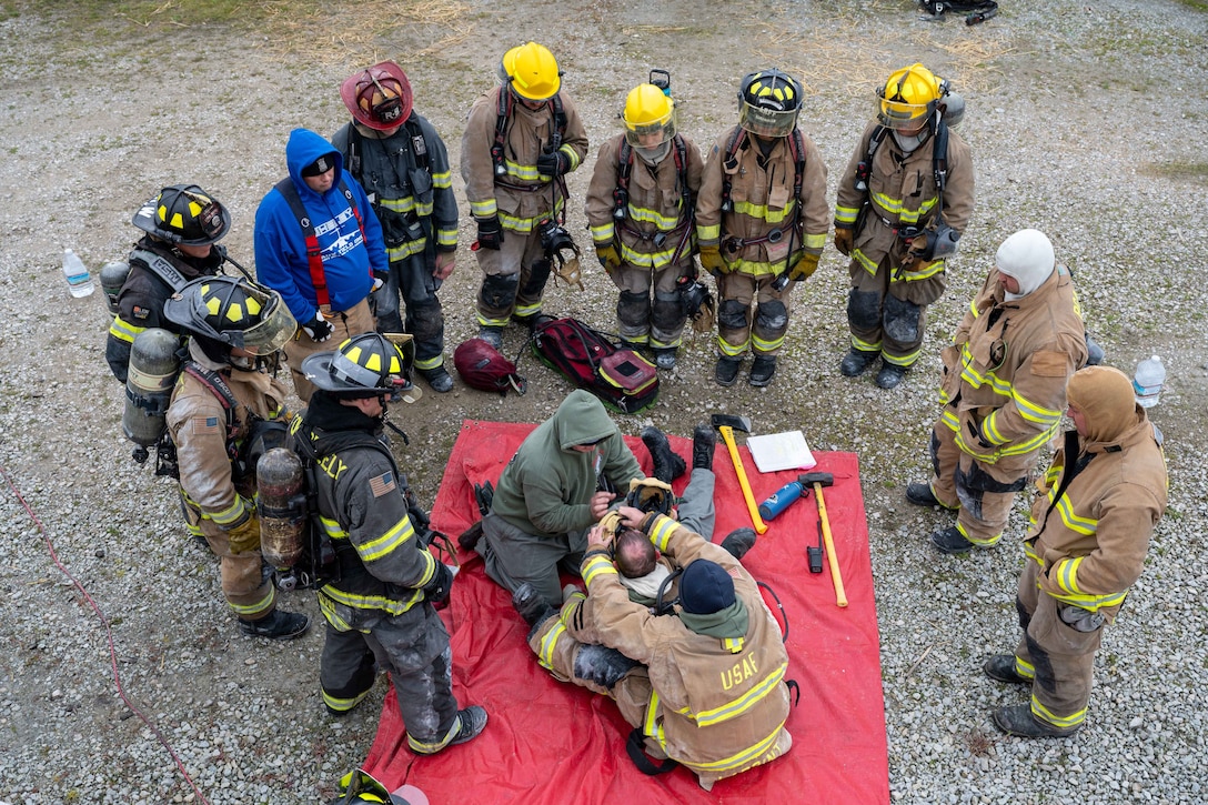Students in firefighting gear stand and watch as three people on the ground demonstrate firefighting procedures on a red tarp. Tools lie around them.