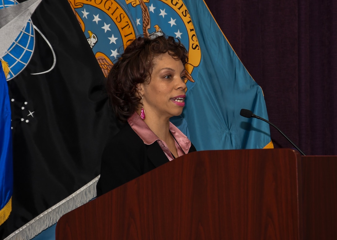Head and shoulders image of Lakeea Kelly standing behind a podium talking into a microphone. the DLA flag is behind her.