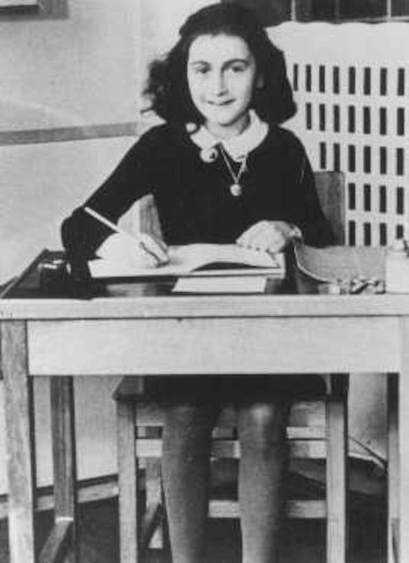 Black and White photo of a young girl behind a school desk. She has a book open and is holding a pencil. She is looking into the camera and smiling
