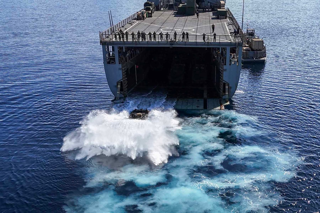 A Marine combat vessel exits a Navy ship and splashes into the water during daylight as service members watch from the flight deck.