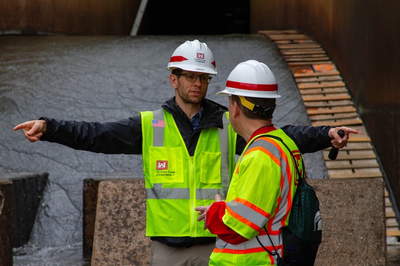 Two men in reflective vests and hard hats talk, one of them pointing with both arms.