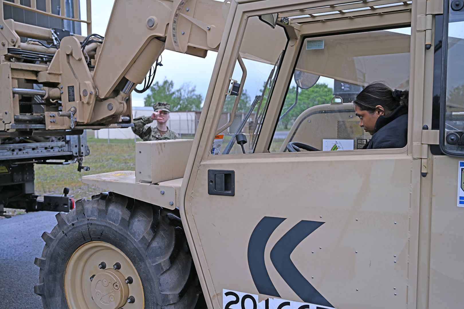 A person in camo uniform directs another person in a camo uniform driving a tan vehicle