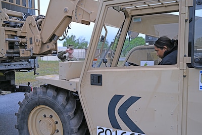 A person in camo uniform directs another person in a camo uniform driving a tan vehicle