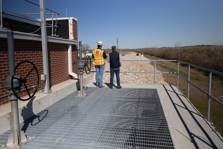 Two men, one in a reflective vest and hardhat, stand at a railing and look at rock with a brick building and metal grates in the foreground.