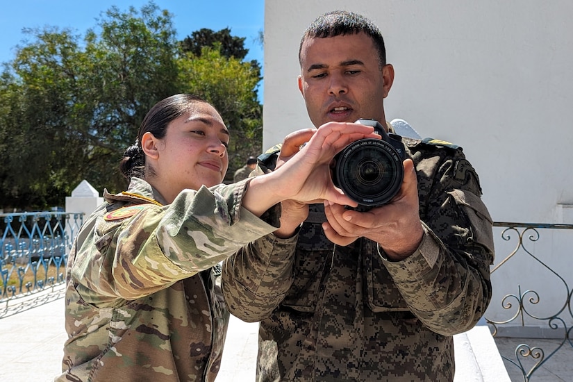 A U.S. soldier adjusts a camera that a foreign service member holds as they stand together outside.