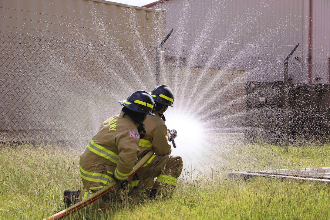 Two soldiers kneel in grass while spraying a hose at a fence with a building in the background.