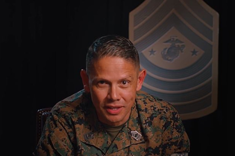 A Marine in uniform speaks against a dark background, with a rank insignia displayed on the wall behind him.