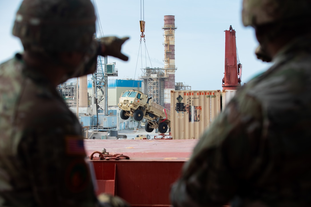 Two soldiers, one pointing, frame a distant image of a military vehicle being hoisted from a shipping container.