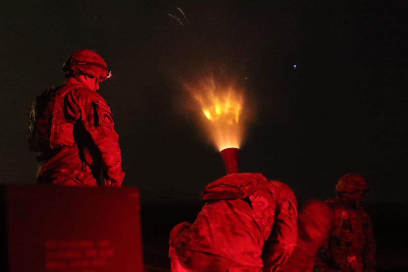 A soldier stands and observes a mortar firing at night to create a bright blaze while others kneel to take cover, all bathed in red light.