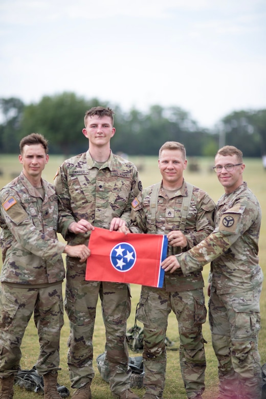 Four soldiers stand in a field jointly holding up a small Tennessee state flag.