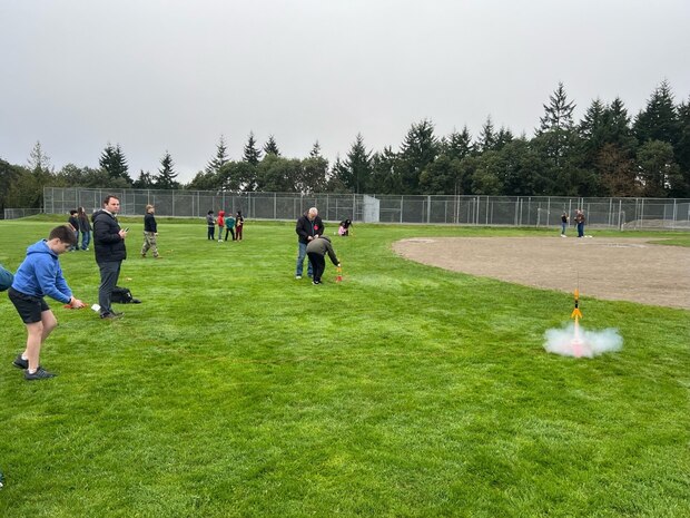 Kevin Durig (left), a Physical Security Specialist at Strategic Weapons Facility-Pacific (SWFPAC) based in Bangor, Washington assists a student from Ridgetop Middle School prepare his rocket to be launched, while another student launches their rocket in the foreground. SWFPAC Science, Technology, Engineering, and Mathematics Volunteers participate in Rocket Launches every school quarter at Ridgetop Middle School.