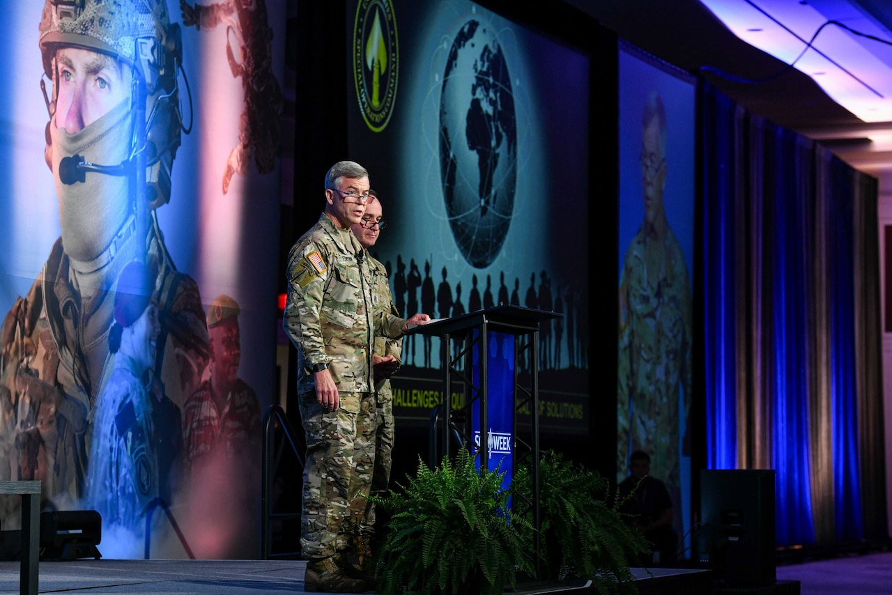 Two service members in camouflage uniforms stand at a lectern speaking to a crowd off screen. “SOF Week” is projected on the wall behind them.