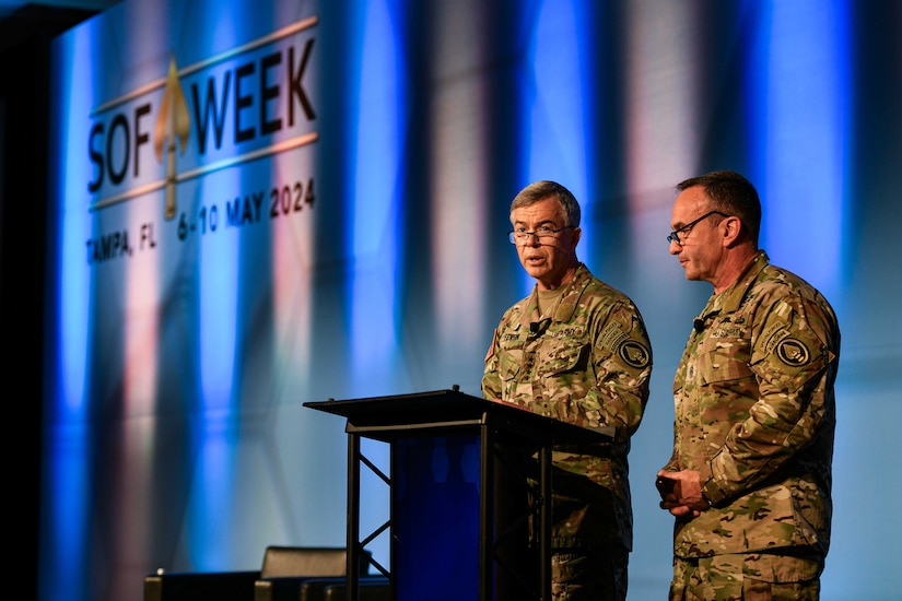 Two service members in camouflage uniforms stand at a lectern speaking to a crowd off screen. “SOF Week” is projected on the wall behind them.