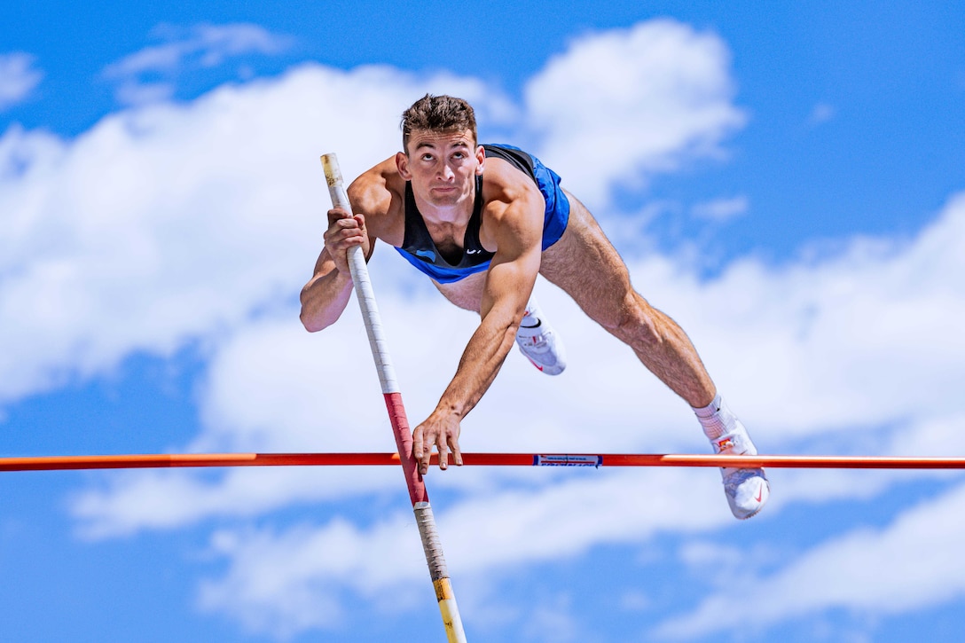 An airman in a track and field uniform looks upward while vaulting over a pole against a partly cloudy sky.