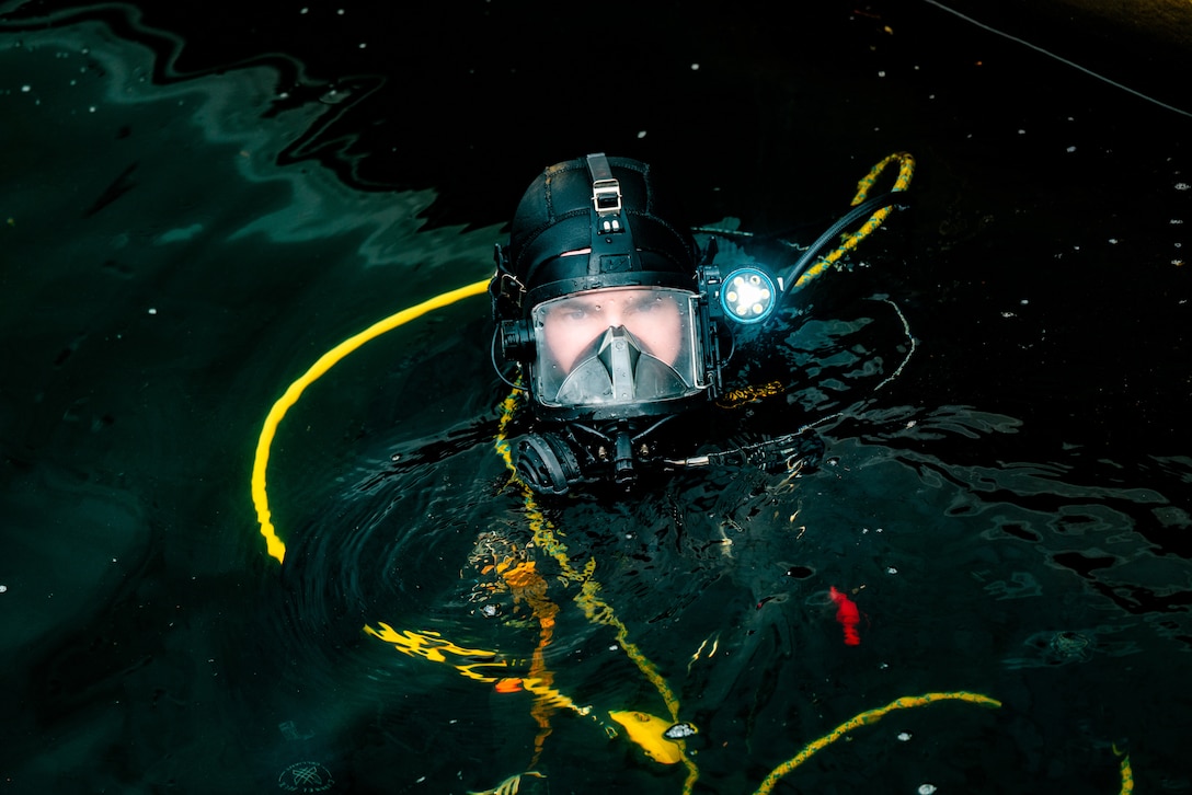 A Coast Guardsman wearing goggles and diving gear looks at the camera while floating in dark water.