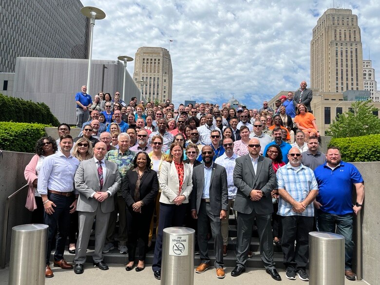 A group of about 200 people stand on steps with tall city buildings in the background and a cloudy sky above.