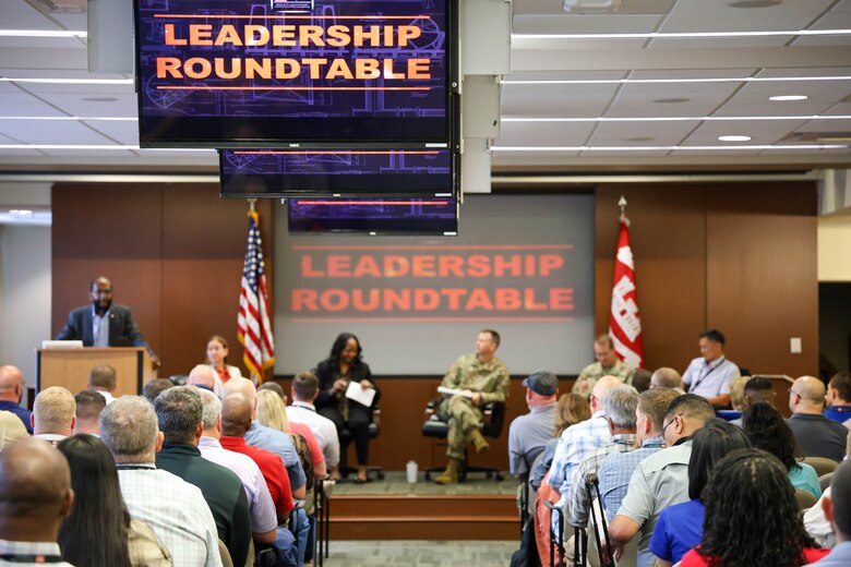 Dozens of people sit in chairs in rows and face a stage where five people sit and one man stands at a podium with the American flag and a projector screen behind him.