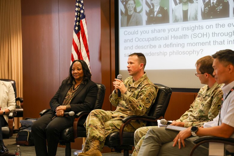 Two women and three men, two of whom are in Army uniforms, sit in a row and one man holds a microphone with the American flag and a projector screen in the background.