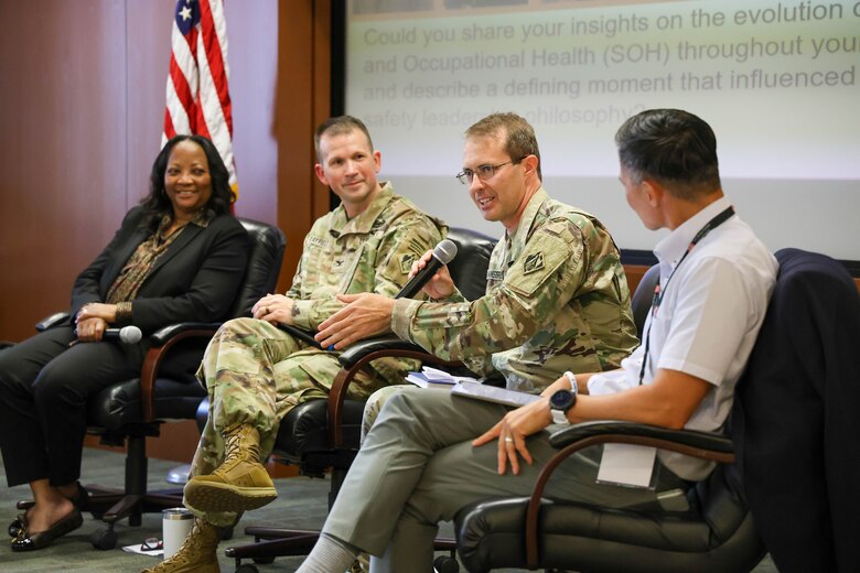 One woman and three men, two of whom are in Army uniforms, sit in a row and one man holds a microphone with the American flag in the background.