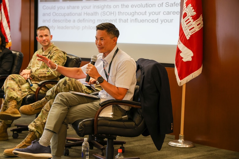 Three men, two of whom are in Army uniforms, and one holds a microphone, sit in a row with a red flag and a projector screen in the background.