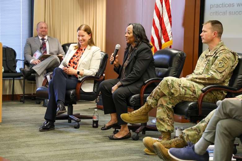 Two women and three men, two of whom are in Army uniforms, sit in a row and one woman holds a microphone with the American flag in the background.