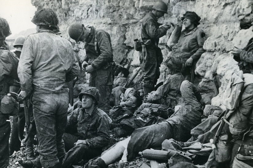 Tired and injured-looking service members rest at the bottom of a cliff face.