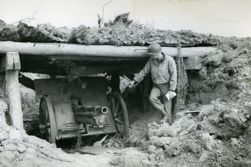 A service member stands beside a large gun hidden inside a cliff.