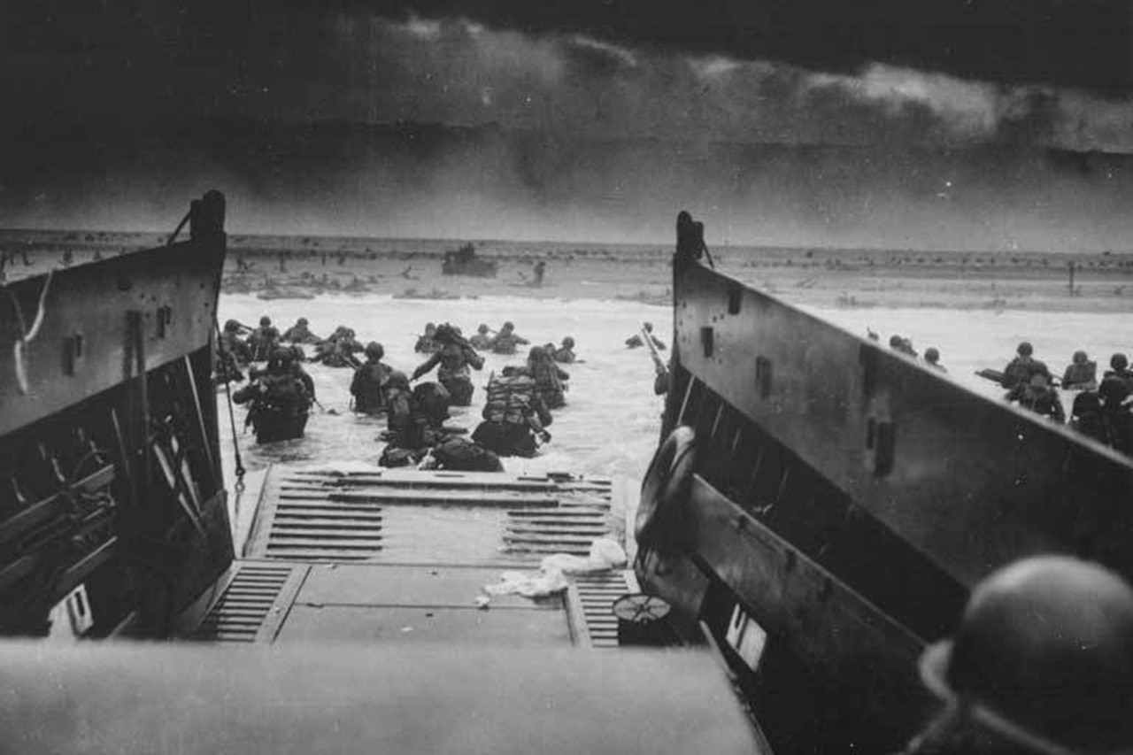 Dozens of service members run into water on a beachhead from a landing craft.