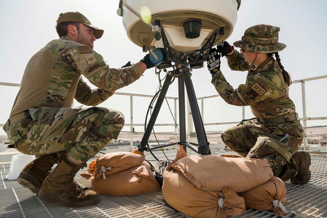 Airmen kneel near sandbags while cleaning a large weather device.