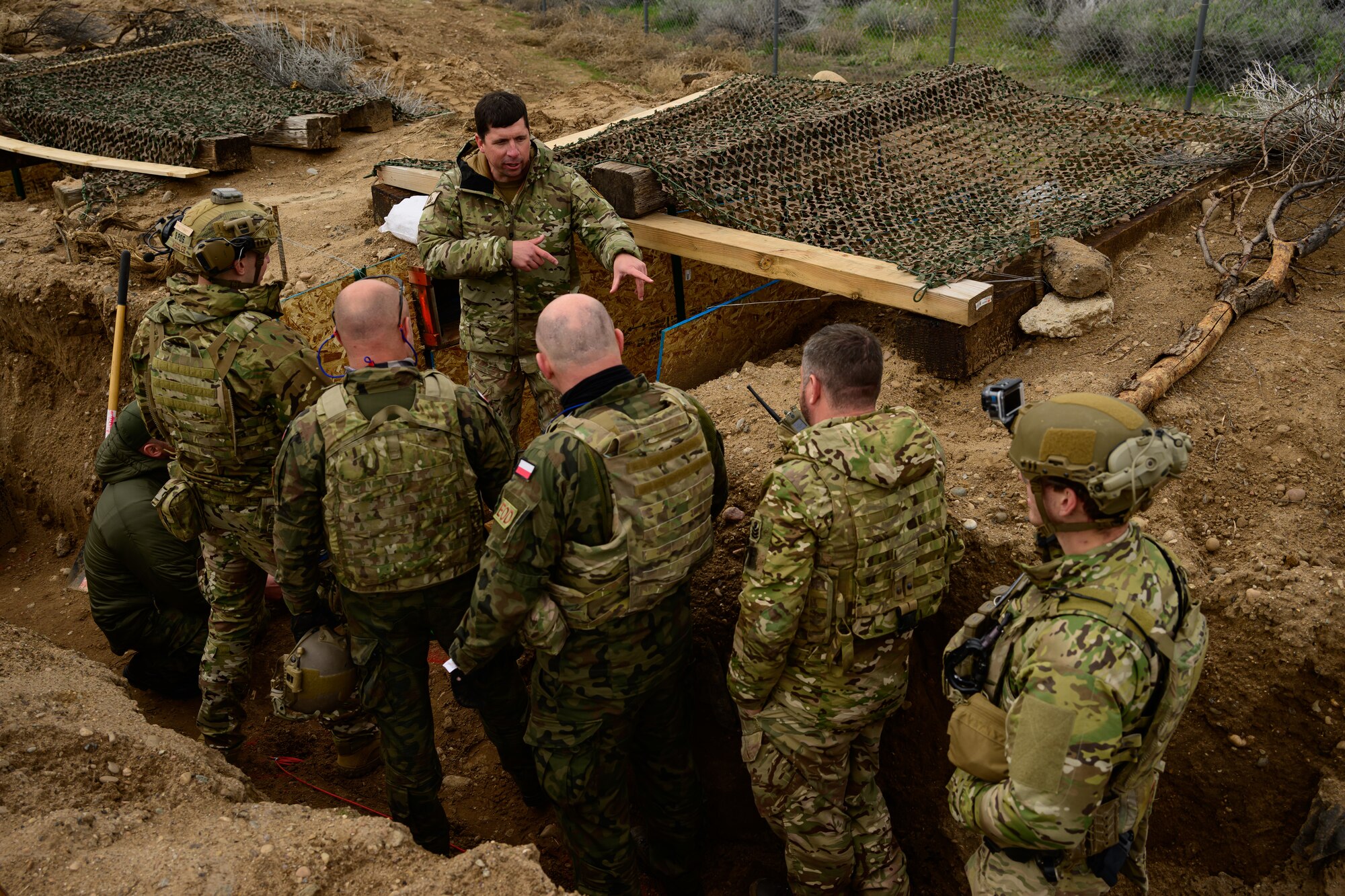 Six men stand in a trench wearing tactical gear.